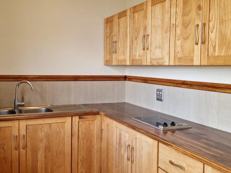 A functional kitchen area featuring wooden cabinetry, a laminate countertop, a stainless steel sink, and a built-in electric hob with tiled backsplash.