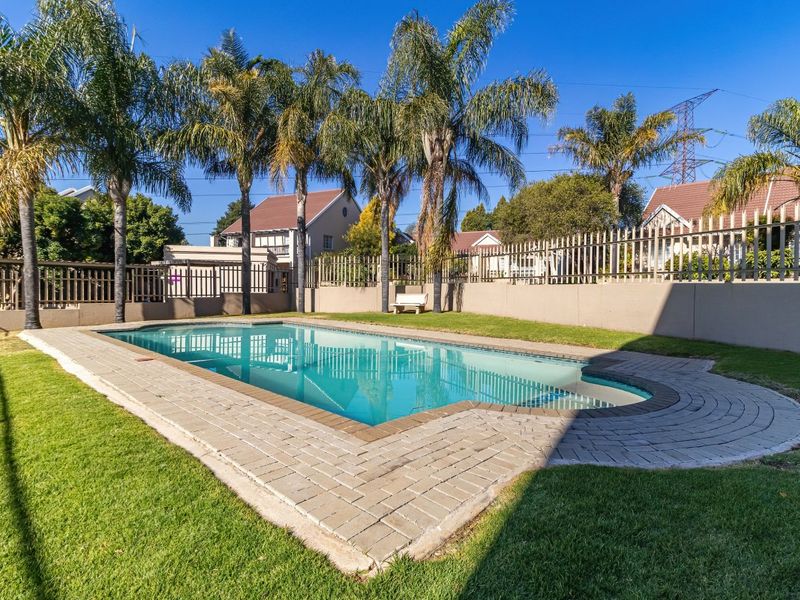 A well-maintained outdoor swimming pool area featuring paved surrounds, manicured lawn, and perimeter walling with metal fencing.