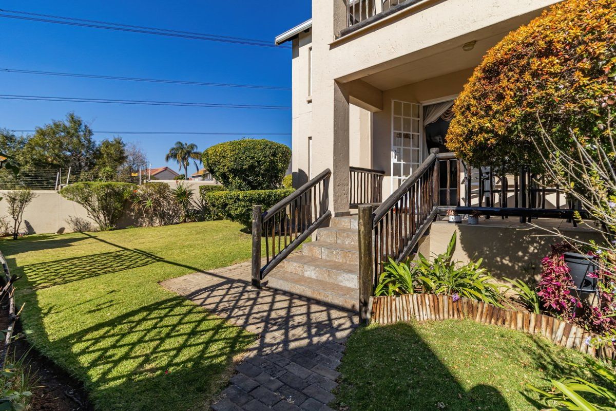 A rear exterior view of the apartment featuring a paved walkway, stone steps leading to a covered patio, and a well-maintained garden area.