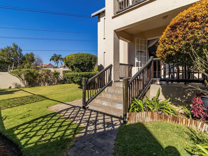 A rear exterior view of the apartment featuring a paved walkway, stone steps leading to a covered patio, and a well-maintained garden area.