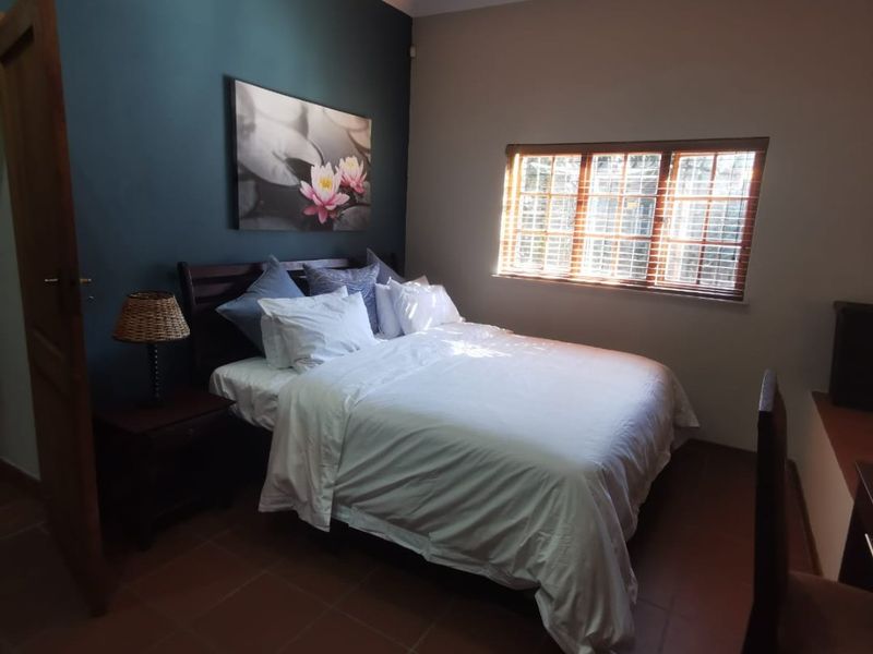 A cozy bedroom featuring terracotta floor tiles, a dark blue accent wall, and natural light filtering through wooden-framed windows with venetian blinds.