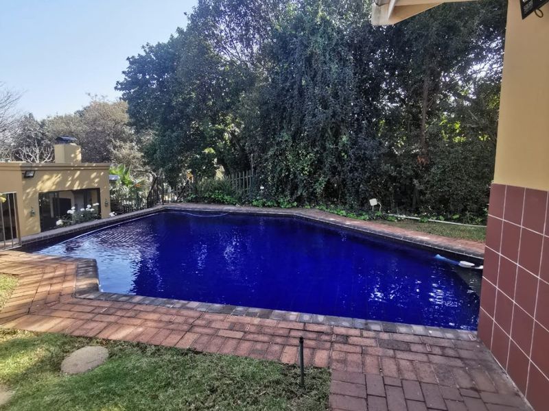 A well-maintained swimming pool area featuring brick paving, a built-in outdoor fireplace, and established garden surroundings.