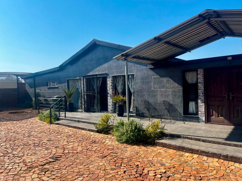 Front exterior view featuring a tiled patio, corrugated metal canopy, and a cobblestone driveway leading to the main entrance.