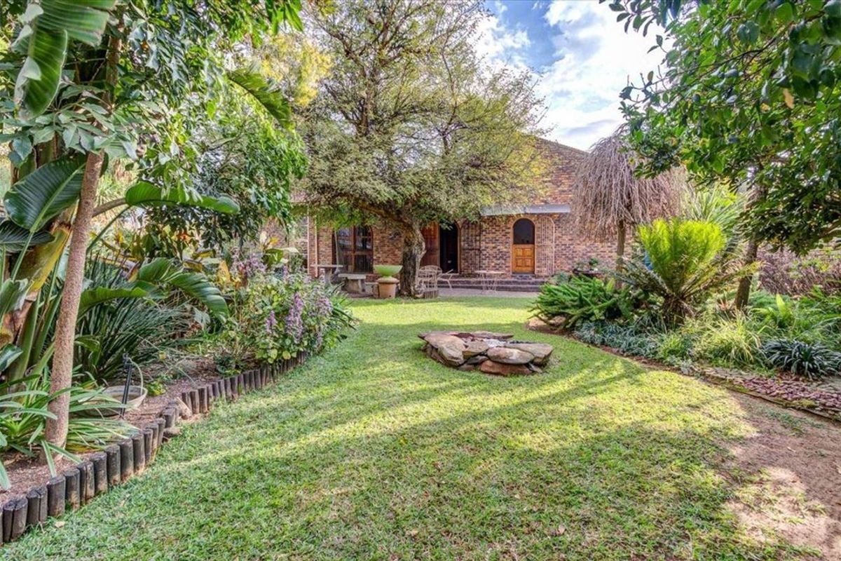 A private rear garden featuring a brick facade, arched entryways, mature trees, and a central stone fire pit area.