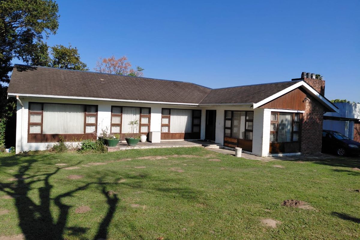 Rear view of a single-story house featuring a pitched roof, brick chimney, and a concrete patio overlooking a spacious lawn.