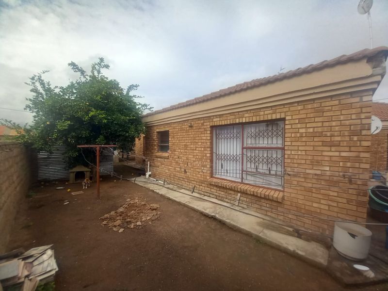 Rear exterior view of a face brick house featuring a tiled roof, security bars on windows, and a surrounding boundary wall with an unpaved yard.