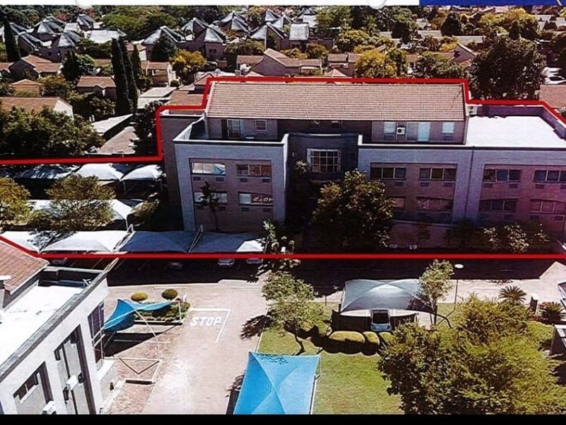 Aerial view of a multi-story commercial office building featuring a tiled roof, paved parking area with shade ports, and surrounding landscaped grounds.