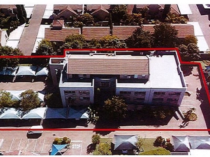 Aerial view of a commercial property featuring a multi-story building, paved parking area with shade cloth carports, and surrounding perimeter boundary.