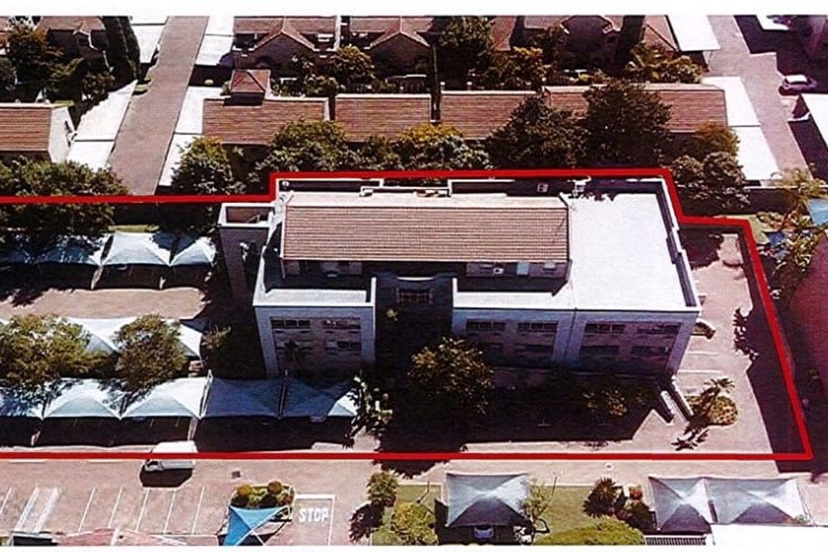 Aerial view of a commercial property featuring a multi-story building, paved parking area with shade cloth carports, and surrounding perimeter boundary.