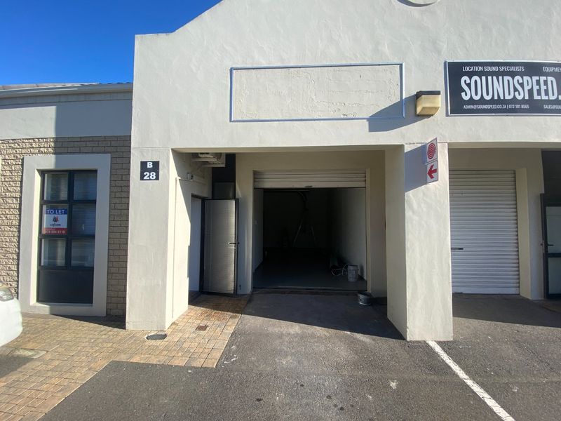 Front elevation of an industrial unit featuring a roller shutter door, pedestrian access gate, and paved driveway area.