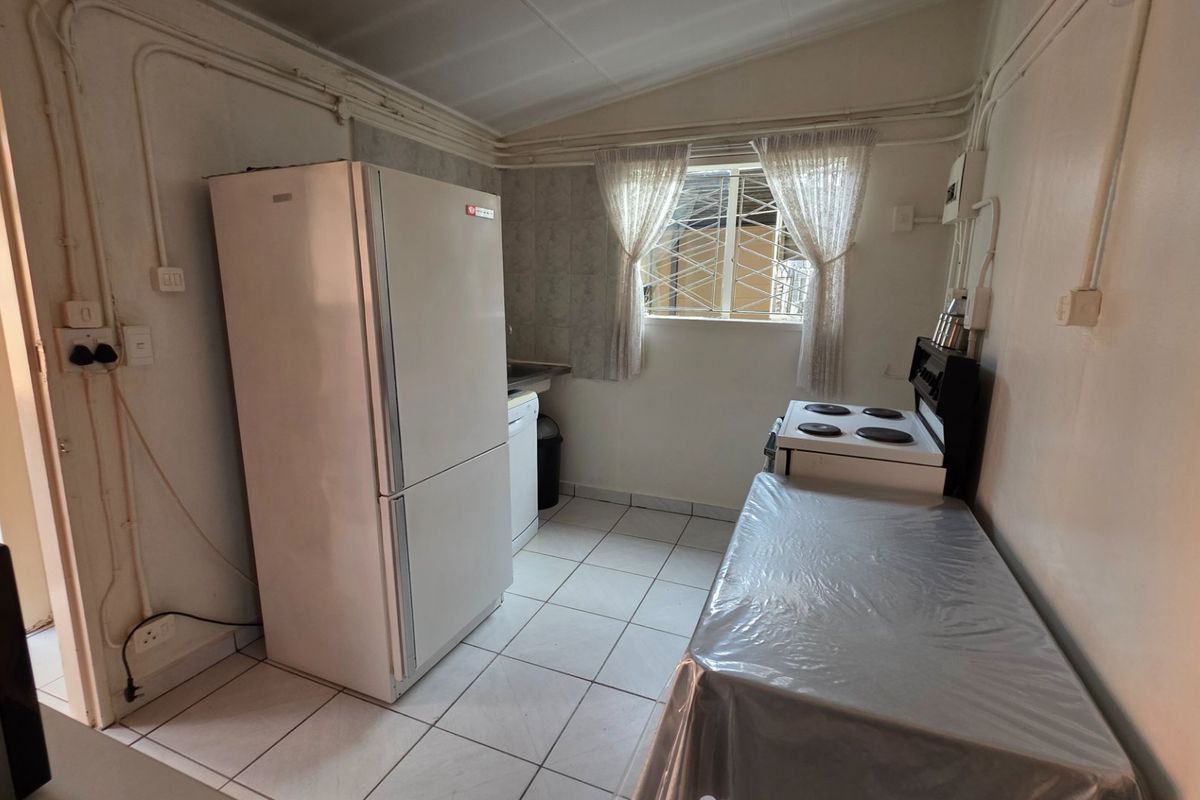 A functional kitchen featuring tiled flooring, an electric stove, and a window with security bars. The space includes exposed electrical conduit and dated finishes.
