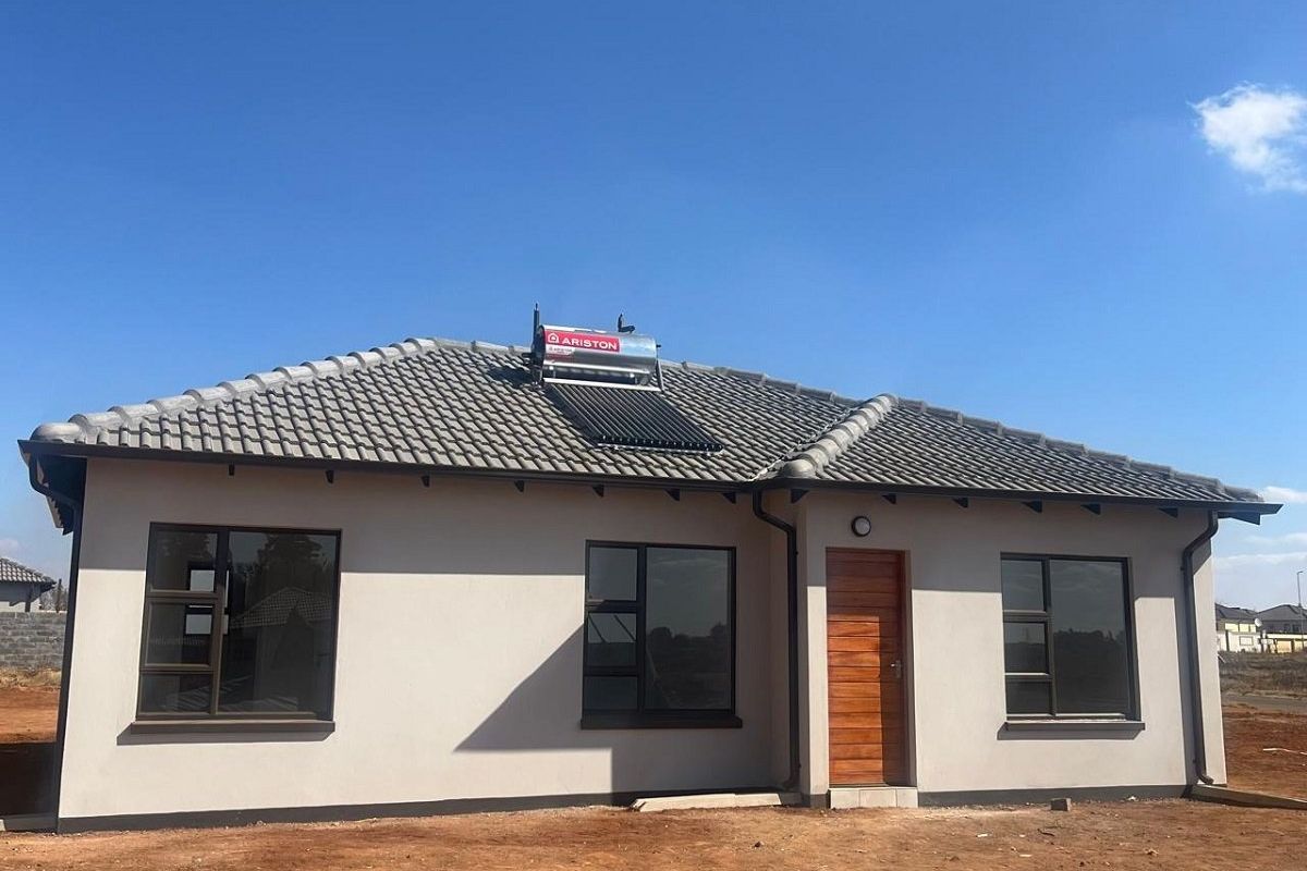 A modern single-story house exterior featuring a tiled roof, solar water heating system, and aluminium window frames, set on an undeveloped plot.