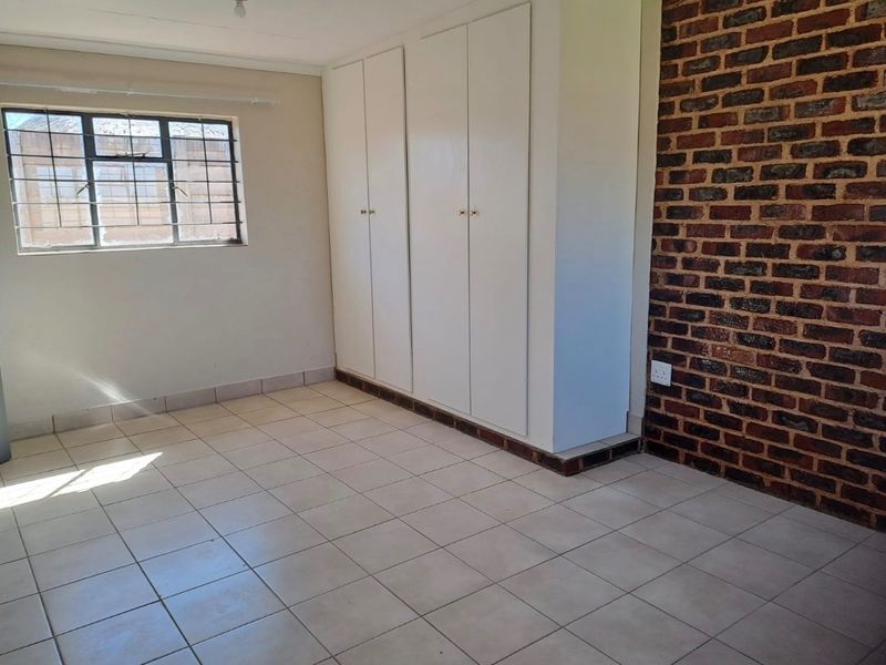 A bright, empty bedroom featuring tiled flooring, built-in cupboards, and an exposed brick feature wall, providing a versatile space for customization.