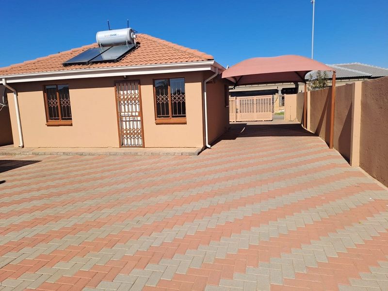 Front view of a well-maintained house featuring a paved driveway, shade sail carport, and solar water heating system on a tiled roof.