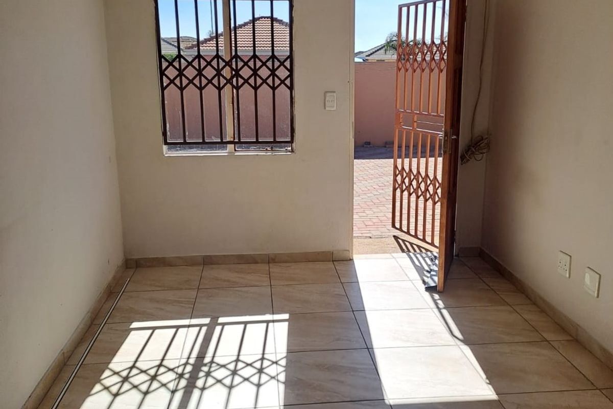 A bright, empty living area featuring ceramic floor tiles, security gates on the door and window, and direct access to the exterior.
