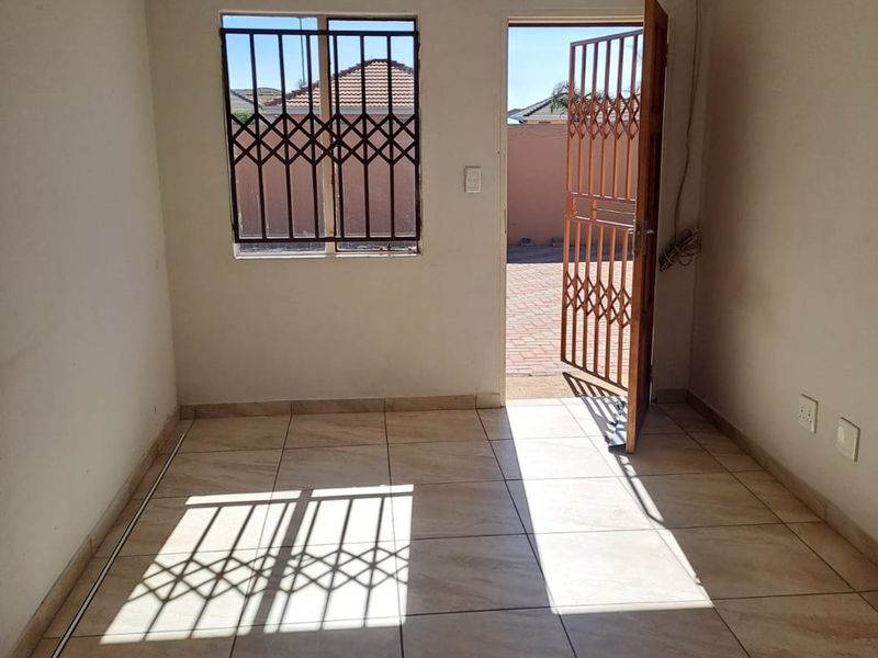 A bright, empty living area featuring ceramic floor tiles, security gates on the door and window, and direct access to the exterior.