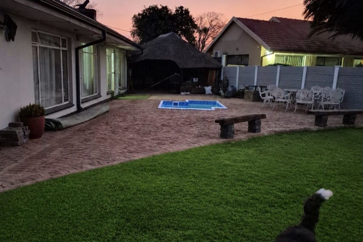 Rear garden area featuring a paved patio, swimming pool, and a thatched lapa structure, set against a sunset backdrop.