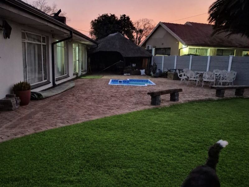 Rear garden area featuring a paved patio, swimming pool, and a thatched lapa structure, set against a sunset backdrop.