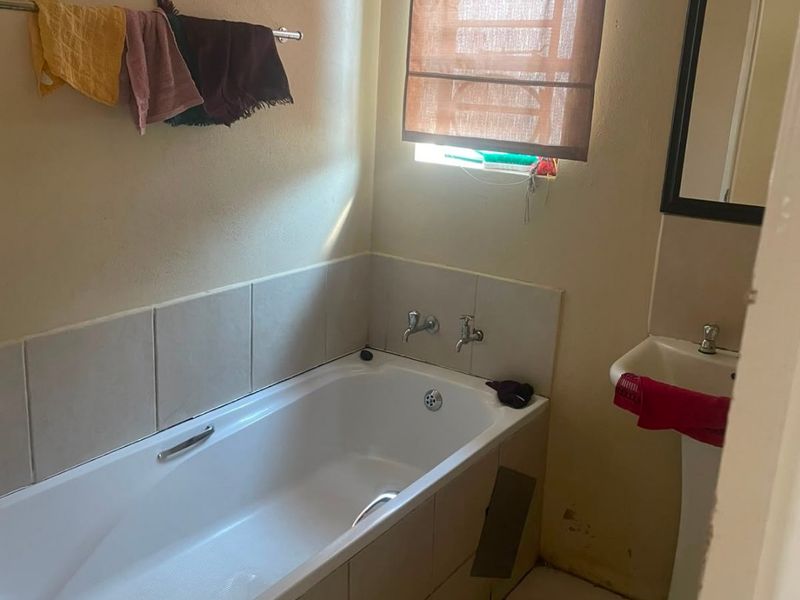 A compact bathroom featuring a bathtub with tiled surround, pedestal basin, and tiled flooring, requiring some maintenance to the wall finishes and bathtub panel.