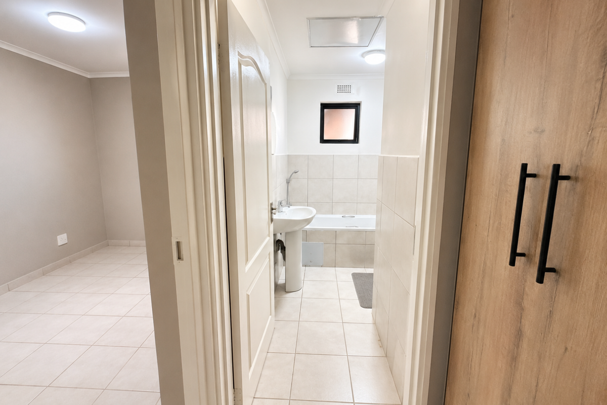 A clean, neutral bathroom featuring a pedestal basin, bathtub, and tiled finishes, viewed from an adjacent room with built-in storage.