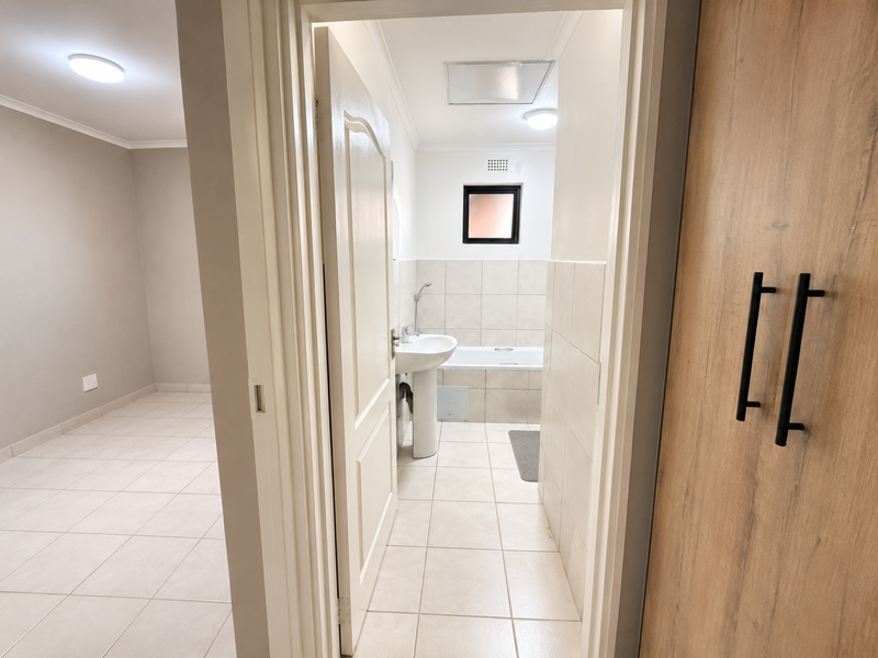 A clean, neutral bathroom featuring a pedestal basin, bathtub, and tiled finishes, viewed from an adjacent room with built-in storage.