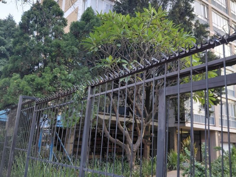 Exterior view of a multi-story apartment building featuring a secure metal perimeter fence with spiked topping and a paved entrance walkway.