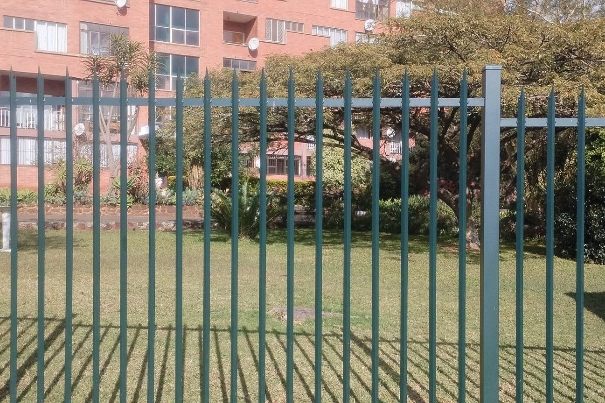 A view of a communal lawn area enclosed by metal spike fencing, with a brick apartment building in the background.