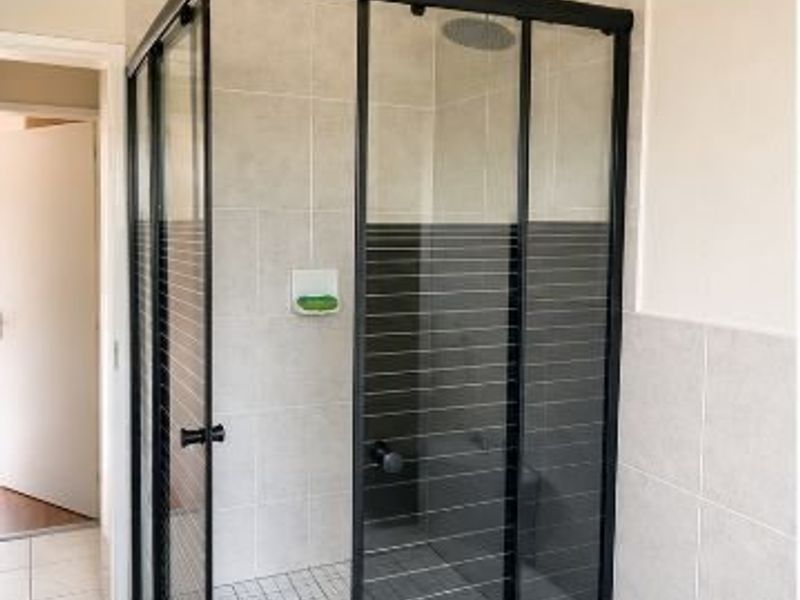 A modern bathroom featuring a corner shower enclosure with black framing, a rain shower head, and neutral wall tiling.