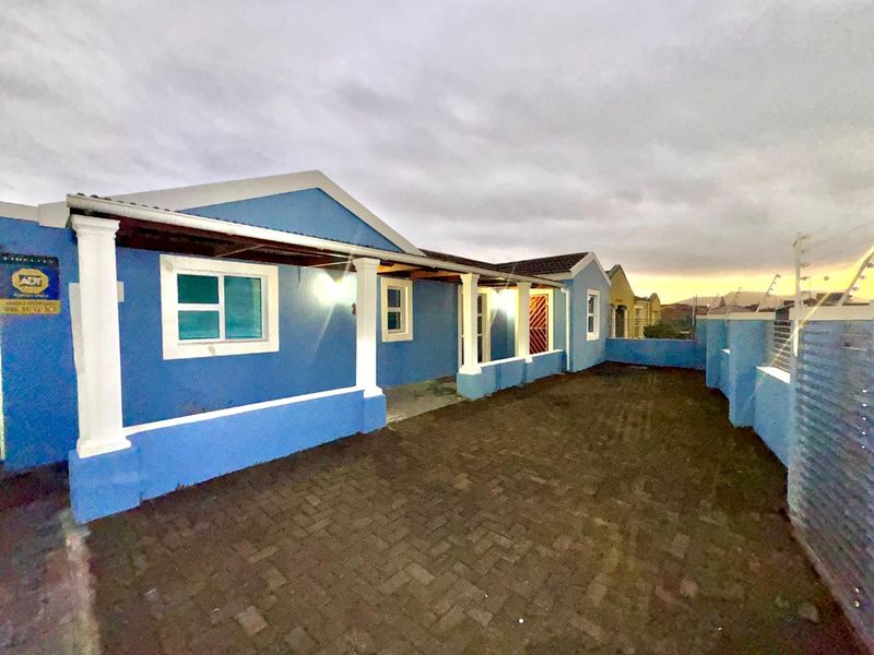 Front exterior view of a blue house featuring a paved driveway, covered porch with columns, and secure boundary wall with electric fencing.