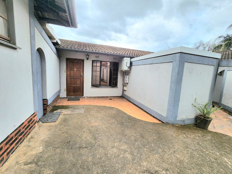 Front entrance area of a townhouse featuring a timber door, tiled patio section, and concrete driveway with visible signs of weathering and wear.