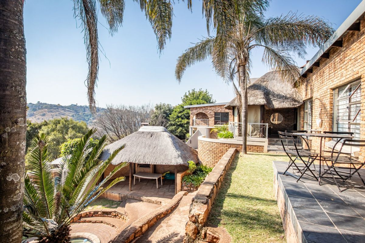 A rear exterior view featuring a thatched lapa, brick architecture, a stone retaining wall, and a paved patio area overlooking the landscape.