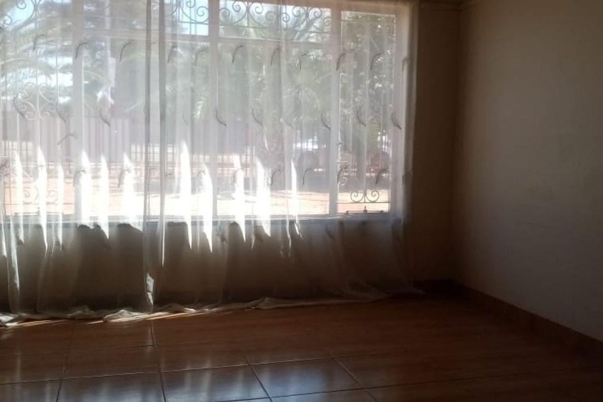 An empty living room featuring tiled flooring and a large window fitted with security bars, allowing natural light into the space.