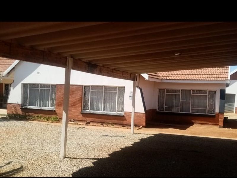 Front exterior view of a single-story house featuring a large carport, brick facade, and gravel driveway area.