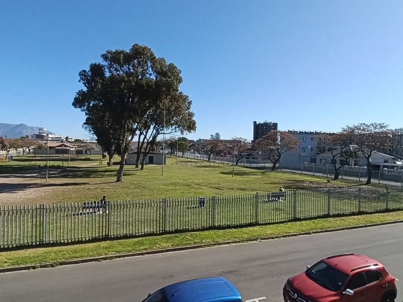 An elevated view overlooking a public park area with mature trees, metal perimeter fencing, and an adjacent street.