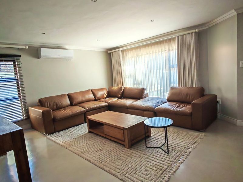A well-maintained living room featuring polished concrete floors, recessed downlighting, and a wall-mounted air conditioning unit with large windows dressed in sheer curtains.