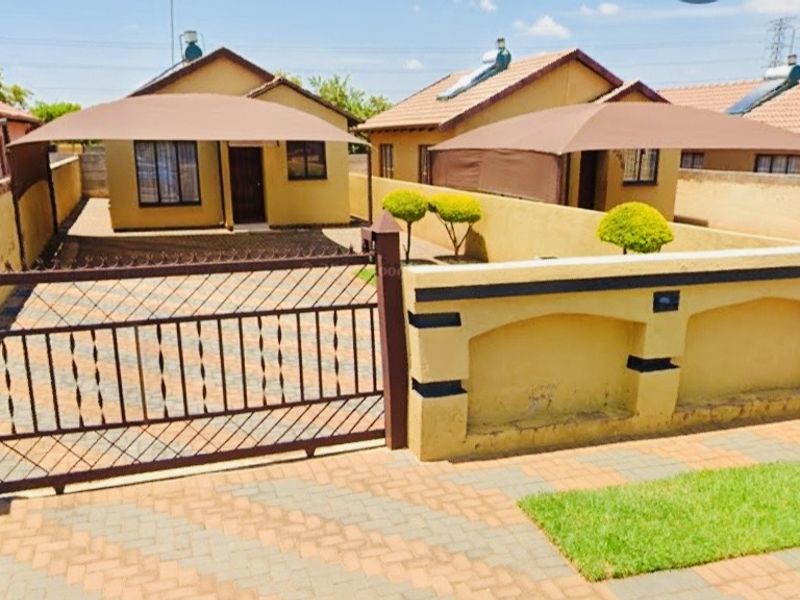 Front view of a residential property featuring a paved driveway, secure metal gate, perimeter wall, and shade sail covering the parking area.