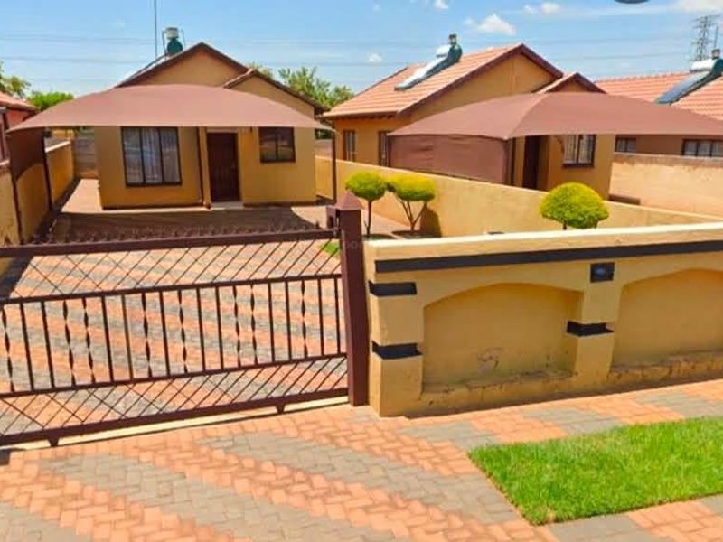 Front view of a residential property featuring a paved driveway, secure perimeter wall with metal gate, and a shade sail over the parking area.
