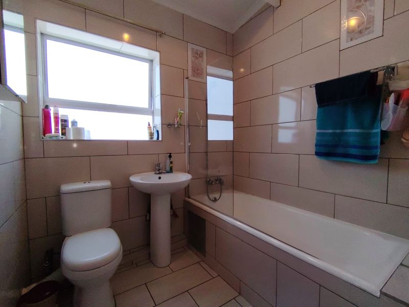 A functional bathroom featuring a pedestal basin, toilet, and a bathtub with a glass shower screen, finished with neutral wall and floor tiling.