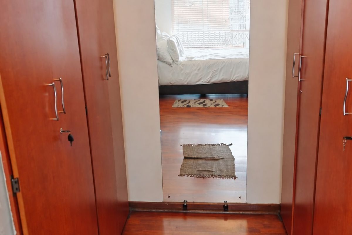 A bedroom dressing area featuring extensive built-in wooden cupboards, laminate flooring, and a central full-length mirror reflecting the sleeping area.