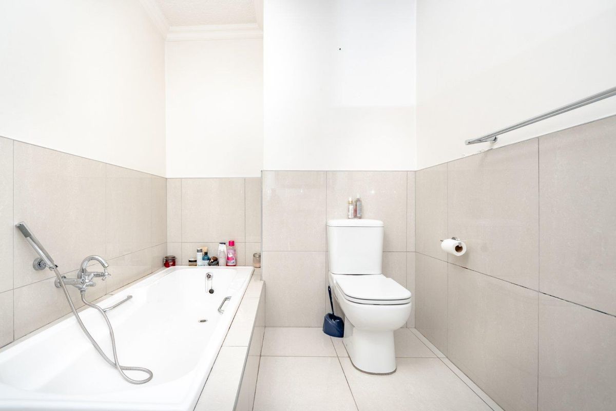 A clean, modern bathroom featuring a built-in bathtub with a handheld shower mixer and a standard toilet, finished with neutral wall and floor tiles.