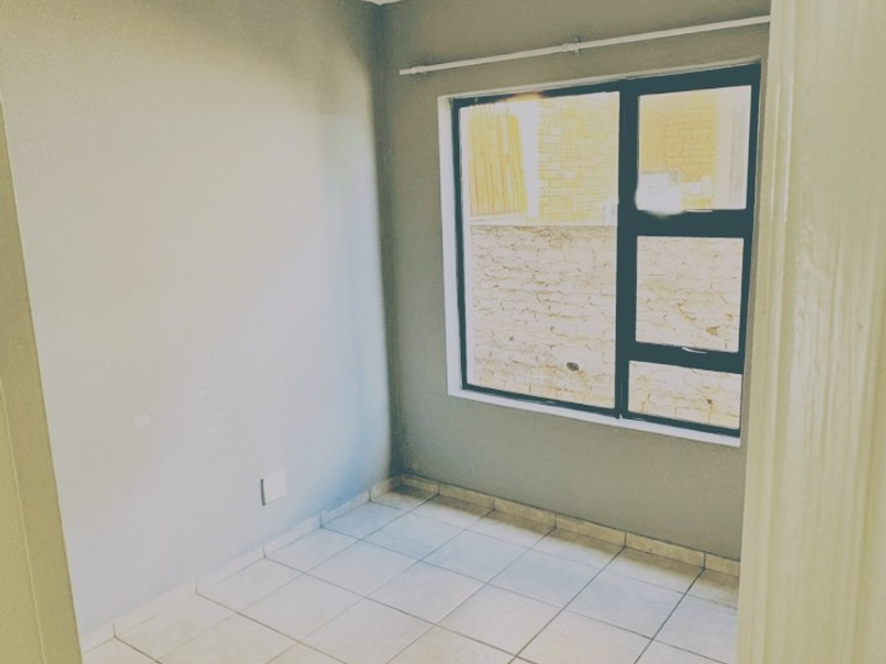 A compact, empty bedroom featuring neutral grey walls, tiled flooring, and an aluminium-framed window with a curtain rail.