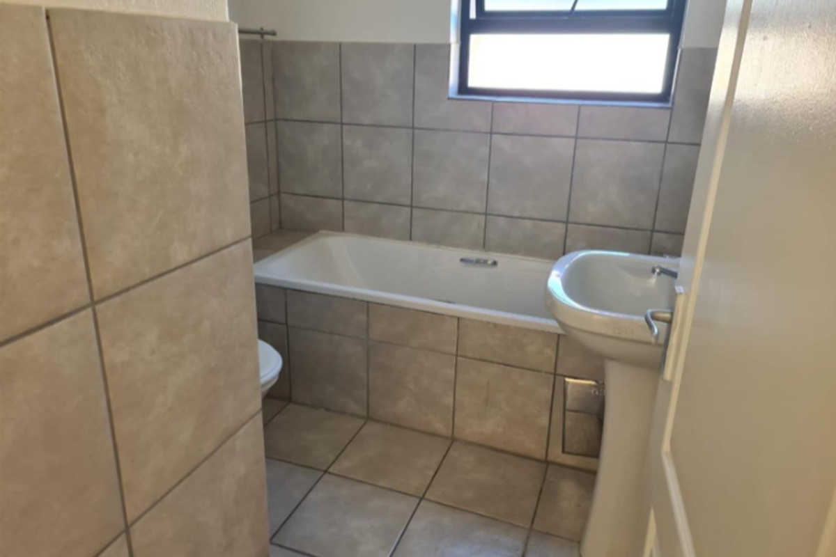 A functional bathroom featuring a bathtub, pedestal basin, toilet, and neutral floor-to-ceiling tiling with a top-hung window for natural light.