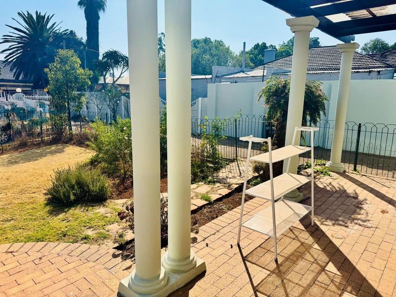 A covered outdoor patio area featuring brick paving and structural columns, overlooking a private garden space.