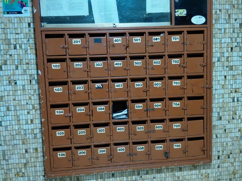 A communal apartment building mailroom area featuring a wall-mounted bank of metal mailboxes and a glass-fronted notice board.