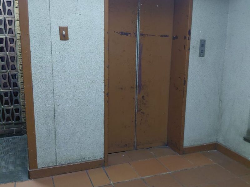 A communal apartment hallway featuring a dated elevator with worn metal doors, ceramic tile flooring, and a decorative breeze block feature wall.