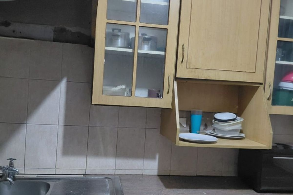 A dated kitchen featuring laminate cabinetry, a stainless steel sink, and tiled backsplash, currently showing signs of wear and missing cabinet hardware.
