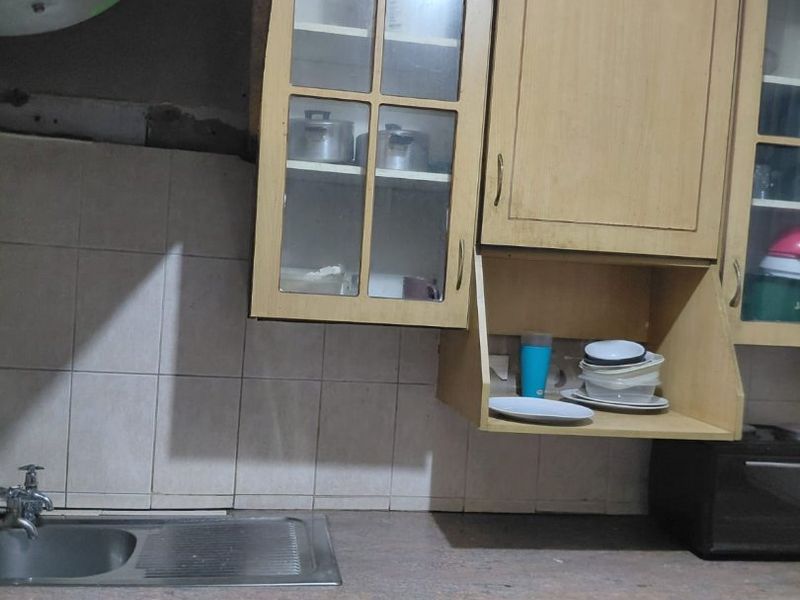 A dated kitchen featuring laminate cabinetry, a stainless steel sink, and tiled backsplash, currently showing signs of wear and missing cabinet hardware.