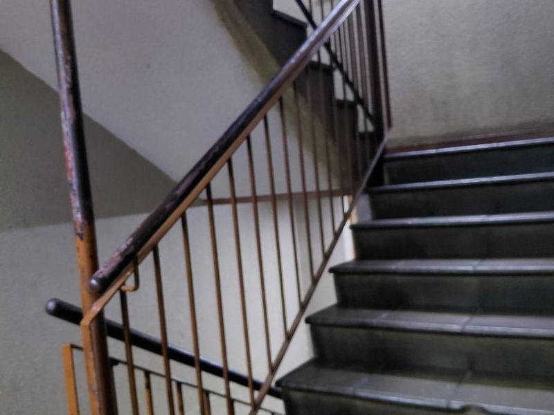 A communal stairwell featuring metal handrails and tiled steps, showing signs of wear and corrosion on the railing.