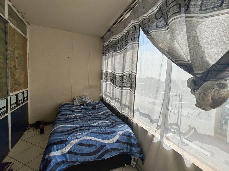 A compact bedroom featuring tiled flooring and a large window with natural light, requiring cosmetic updates to the wall finishes.