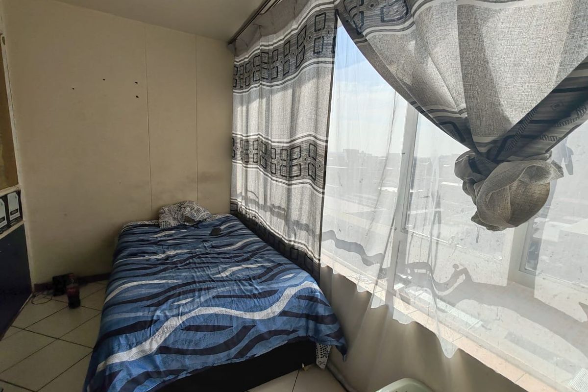 A compact bedroom featuring tiled flooring and a large window that provides ample natural light, currently requiring minor cosmetic wall repairs.